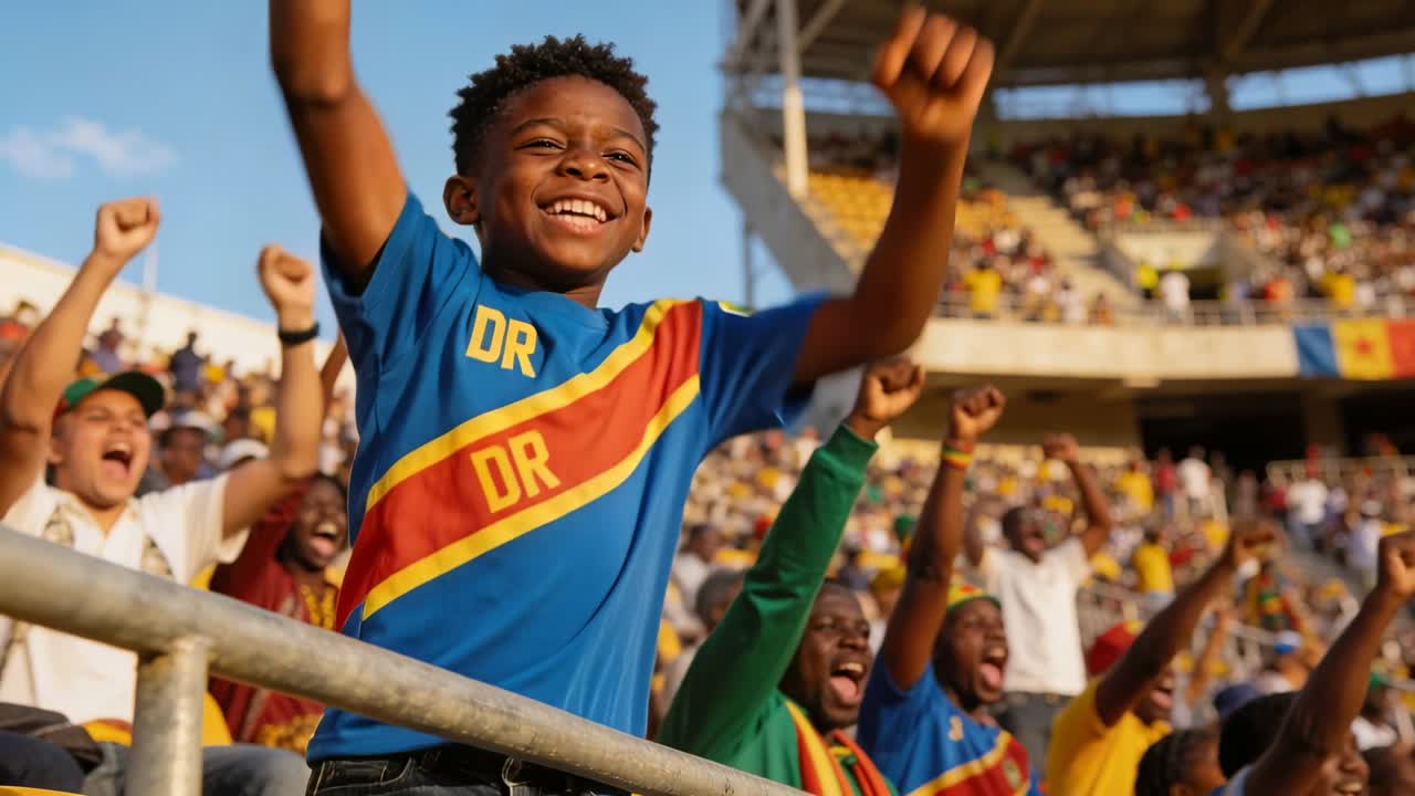 Cheering boy wearing blue DR shirt lifting arms at stadium railing after play, fans pumping fists