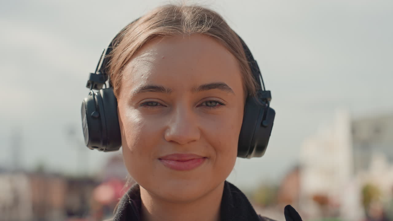 Ung kvinde med hovedtelefoner smiler direkte ind i kameraet i et pulserende gadebillede, blødt sollys i ansigtet, selvsikkert blik, nærbilledeportræt, naturlig makeup, afslappet jakke, subtile bevægelser, venlig
