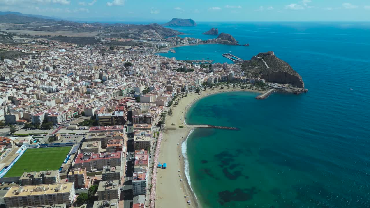 Slow backward aerial pullback over Águilas in Murcia, Spain, revealing the coastline, beaches, harbour area and the Estadio El Rubial football stadium on the left side of the city