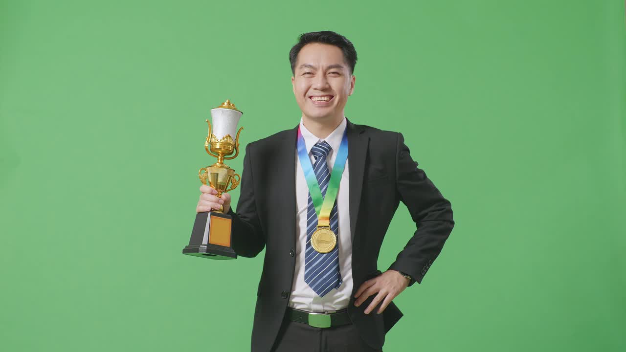 슈트와 타이를 입은 아시아 사업가, 금메달과 트로피를 들고, 한 팔로 카메라를 향해 미소 짓고, 스튜디오의 녹색 스크린 배경에서 첫 번째 우승자로 포즈를 취하고 있다.