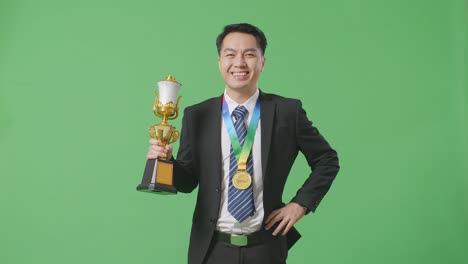 hombre de negocios asiático en traje y corbata con una medalla de oro y un trofeo sonriendo a la cámara con un brazo akimbo posando como el primer ganador en el fondo de la pantalla verde en el estudio