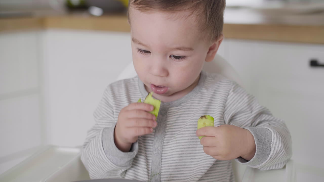 Primo piano di un bimbo carino che mangia fettine di avocado seduto sul seggiolone in cucina