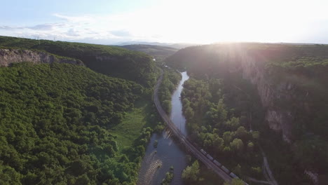 Vorbei-An-Einem-Zug-Auf-Einer-Brücke-über-Einen-Fluss-Bei-Sonnenuntergang,-Umgeben-Von-Wald---Antenne