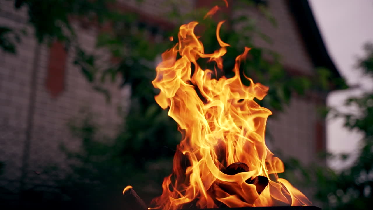 Smoldered logs burned in vivid fire on the house background. Fantastic orange and yellow flame of campfire outdoors. Close-up.