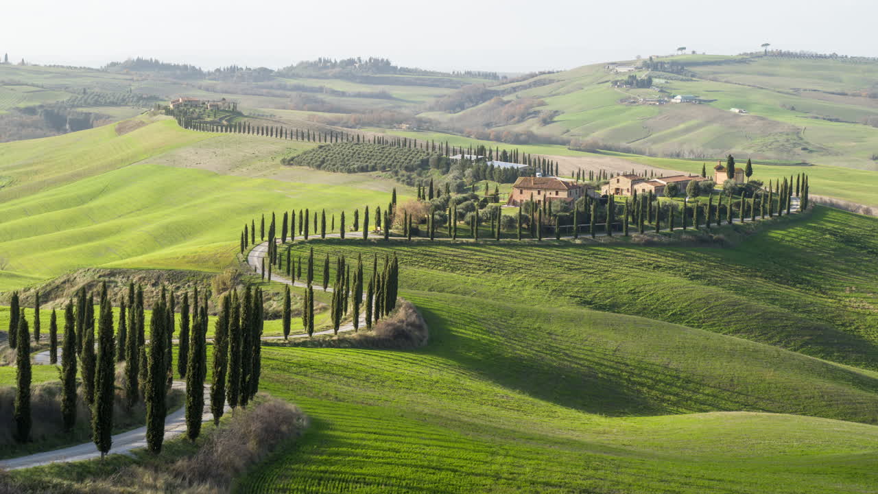 A breathtaking aerial view of the iconic Tuscan countryside in Italy, showcasing rolling green hills, a winding cypress-lined road, and a traditional farmhouse estate. The tranquil rural setting.