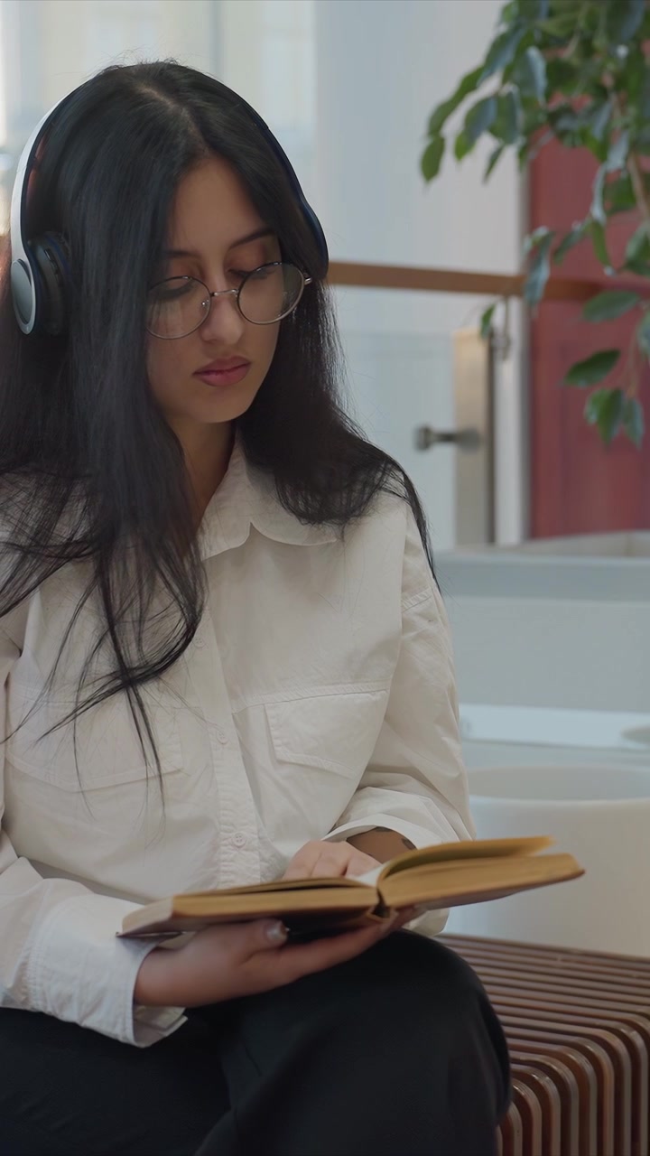 Primer plano de una mujer joven con cabello largo y oscuro usando gafas. La mujer viste una camisa blanca abotonada sobre pantalones negros. La escena muestra un movimiento mínimo mientras la mujer gira ligeramente su cuerpo