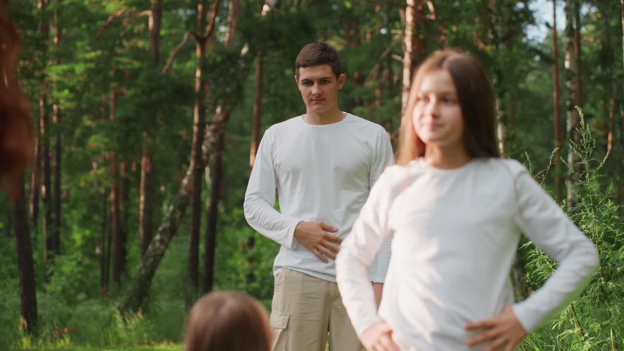 Young girl standing in front while big brother stands behind her in lush green forest under warm sunlight, creating peaceful family outdoor atmosphere filled with quiet emotion and natural beauty