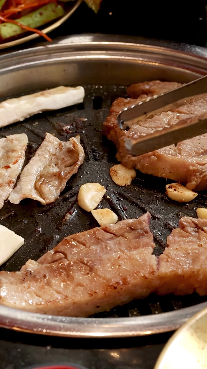 Irisan daging babi marinasi dan bawang putih dipanggang di barbekyu meja, penjepit membalik daging, pencahayaan hangat.