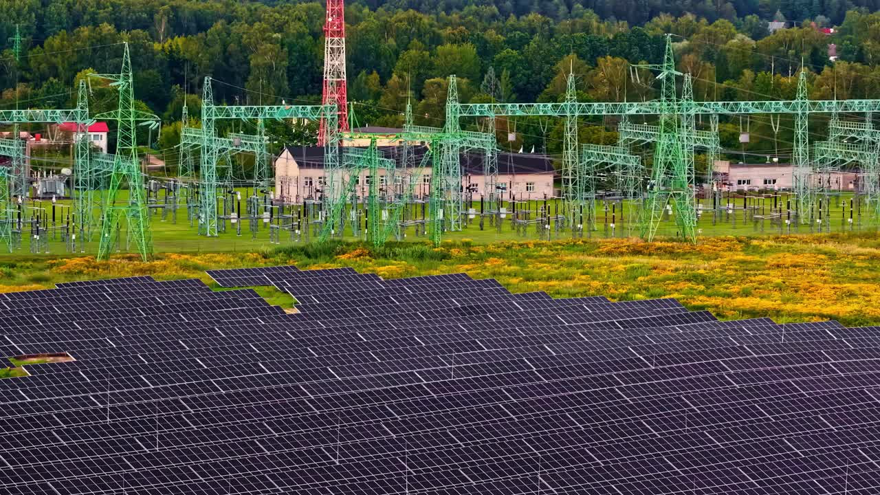 Low aerial glide over large solar panel array directly beside green high-voltage electricity substation with pylons, transformers, red tower and forest backdrop