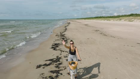 aerial view with a young longhaired girl riding a bike on the sandy beach, waving hand, sunny day, white sand beach, active lifestyle concept, wide drone shot moving backward