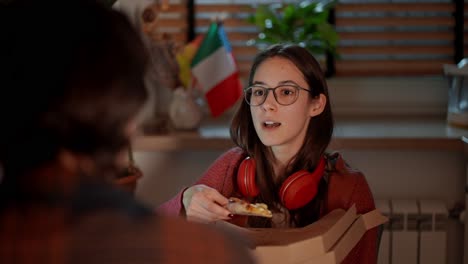 Por-Encima-Del-Hombro,-Una-Chica-Morena-Segura-De-Sí-Misma-Con-Gafas-Y-Auriculares-Inalámbricos-Rojos-Come-Pizza-Durante-Su-Merienda-Mientras-Estudia-Idiomas-Extranjeros-Con-Su-Profesora-Morena-En-Un-Moderno-Apartamento-En-La-Cocina-Cerca-De-Las-Banderas-De-Italia.