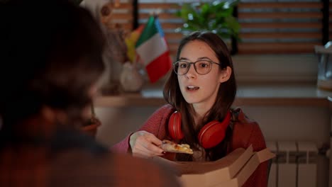 sobre el hombro una confiada chica morena con gafas y auriculares inalámbricos rojos come pizza durante su bocadillo mientras estudia lenguas extranjeras con su maestra morena en un apartamento moderno en la cocina cerca de las banderas de italia