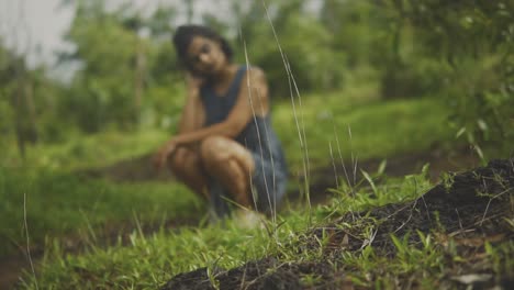 Woman-squats-on-hillside-with-head-resting-on-arm,-rack-focus-to-blades-of-grass