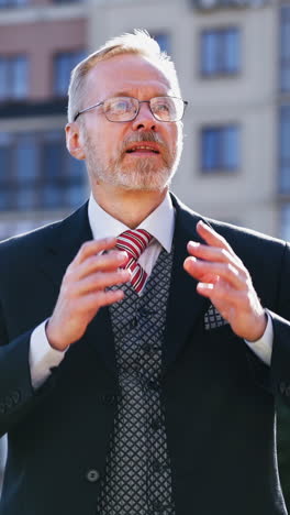 portrait senior businessman in eyeglasses on the building background in the city. serious bearded man with grey hair in suit telling about his business outdoors. vertical video