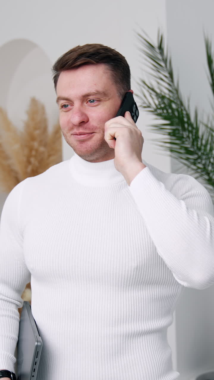 Cheerful young man wearing white sweater speaks on the phone. Businessman with laptop in hands looks at camera. Vertical video
