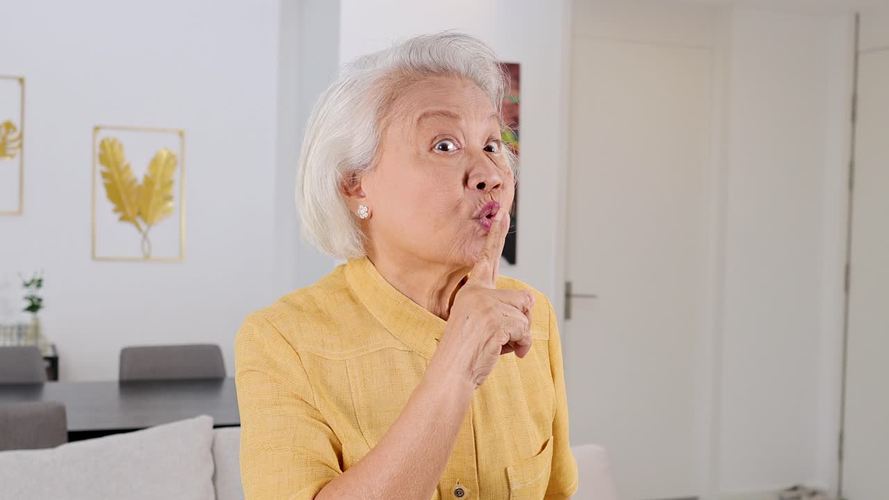 Senior woman in yellow shirt signals silence indoors, bright lighting, steady camera, neutral mood