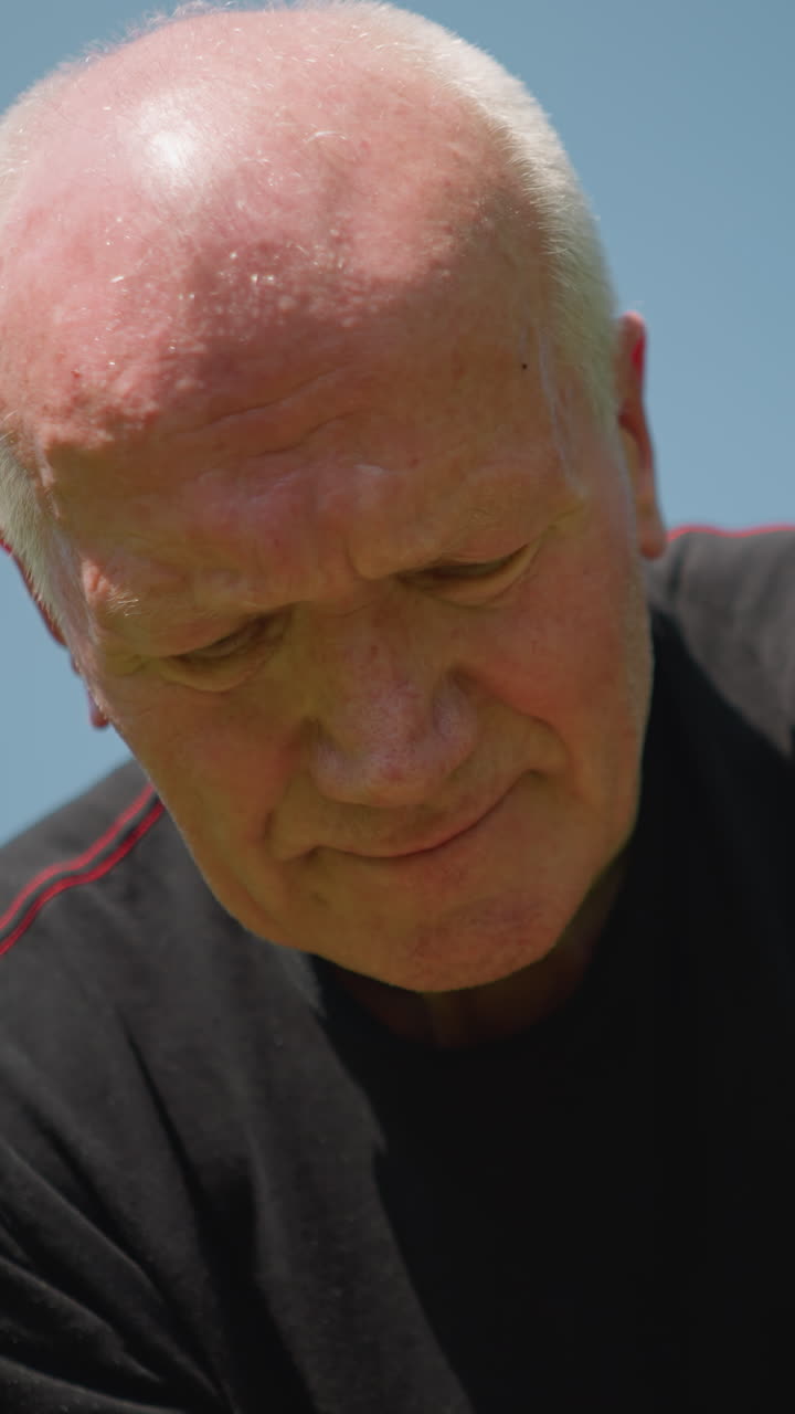 An elderly man is kneeling down, deeply focused on working on something as he engages in his task with a blurred view of trees in the background