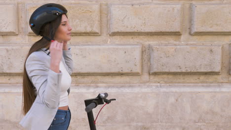 smiling young woman putting on helmet and riding electric scooter in the city