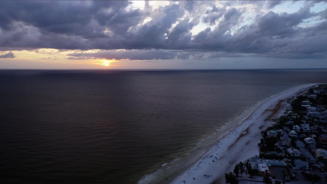 Linia brzegowa Anna Maria Island delikatnie lśni, gdy słońce zachodzi za gęstymi chmurami o zmierzchu, rzucając ciepłe refleksy na spokojne wody Gulf obok cichych domów na plaży.