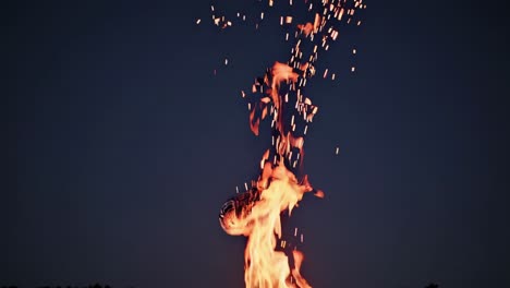 bright orange and yellow flames rising from a campfire with sparks flying up against a dark blue twilight sky, creating a mesmerizing and warm ambiance