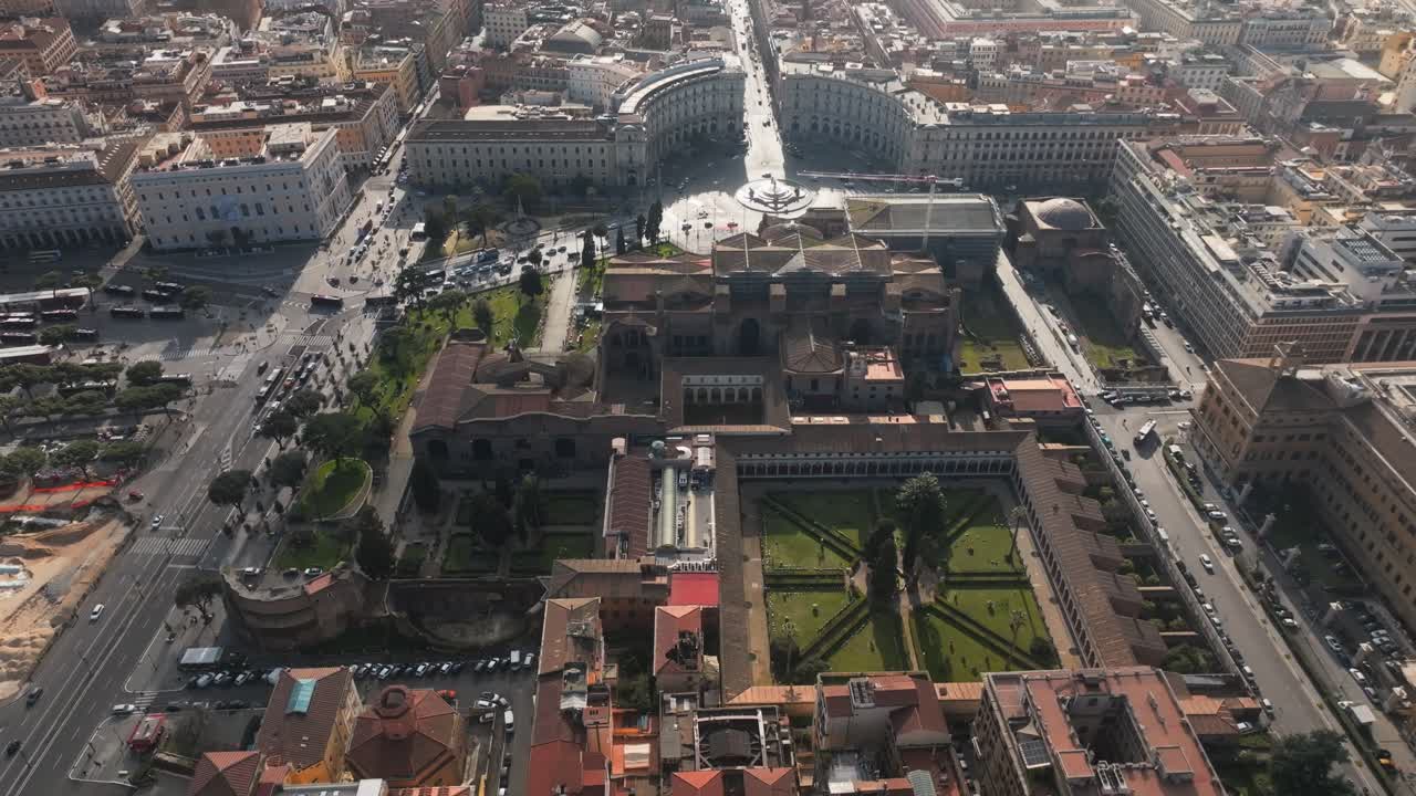 Ripresa aerea cinematografica che rivela l'architettura storica di Roma, con un giardino a chiostro simmetrico e il paesaggio urbano sotto un cielo luminoso.