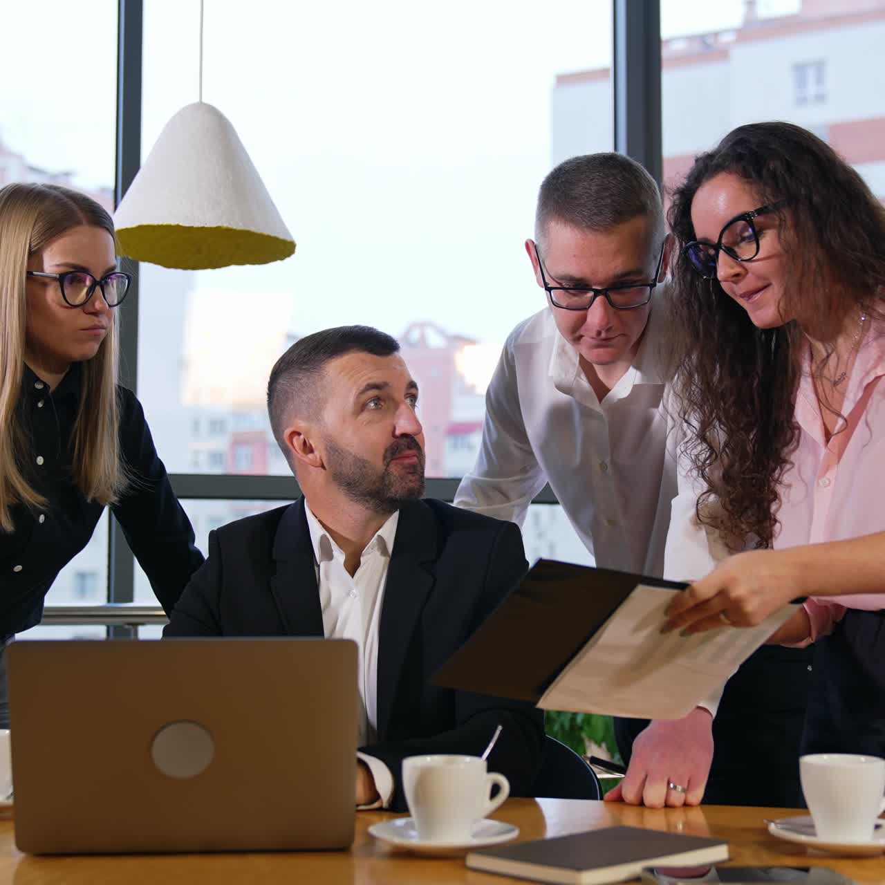 Strict serious boss sitting at the desk with laptop is ready to hear the report. Colleagues gathered around him to work out new ideas