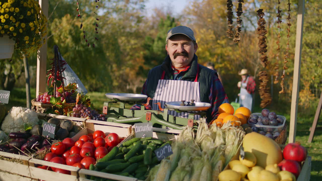Senior Farmer Stands at the Stall and Looks at Camera Senior Farmer Stands at the Stall with Fresh Colorful Fruits and Vegetables Looks at Camera Weekend Shopping at Local Farmers Market Outside Vegetarian Organic and Healthy Food Agriculture