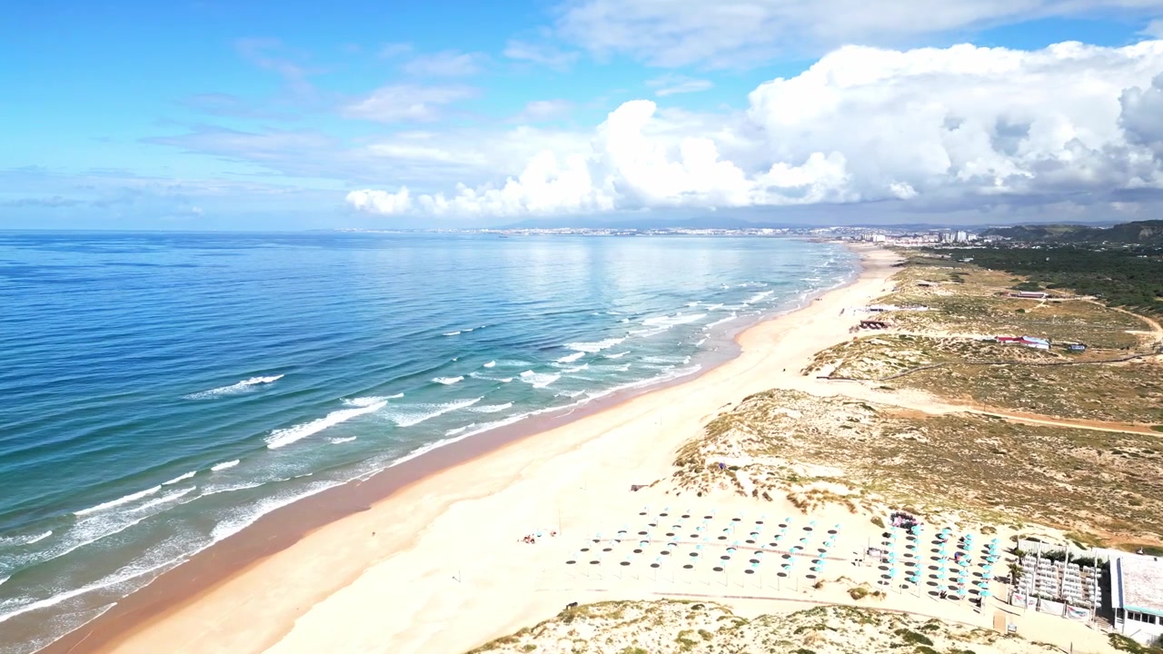 Vista aérea de uma longa praia de areia com ondas e guarda-sóis, céu azul límpido acima