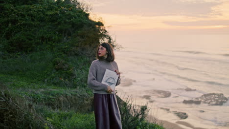 Woman-looking-overcast-seashore-nature.-Girl-artist-holding-album-with-drawings.