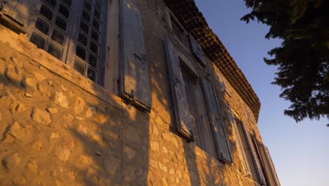 Pared-De-Una-Antigua-Casa-De-Pueblo-De-Piedra-Francesa-Al-Atardecer-Hora-Dorada-Con-Ventanas-Y-árboles-En-El-Callejón