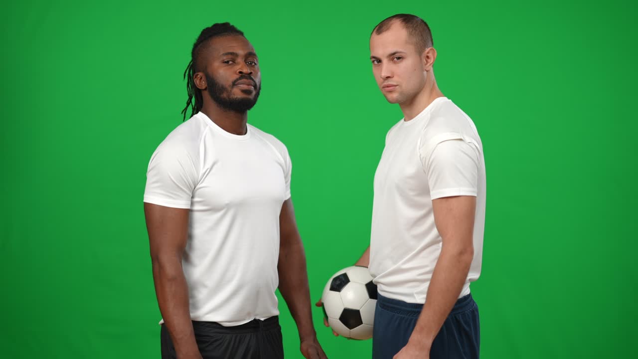 Serious motivated African American and Caucasian men standing at chromakey background face to face turning looking at camera. Portrait of concentrated sportsmen with soccer ball on green screen.