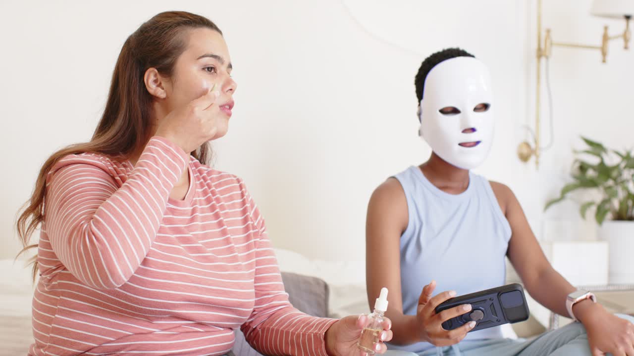 Afroamerikanske kvinder påfører serumdråber og bruger fjernbetjening til LED-maske i sofaen til ansigtspleje.