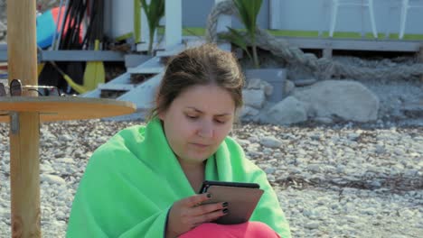 Una-Turista-Envuelta-En-Tela-Verde-Leyendo-Usando-Su-Tableta-Digital-Mientras-Se-Sienta-En-La-Orilla-De-Guijarros-En-La-Playa-De-Agia-Kiriaki,-Cefalonia,-Grecia