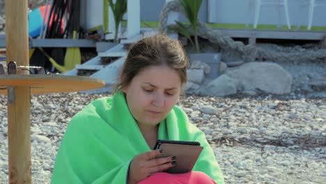 una turista envuelta en tela verde leyendo usando su tableta digital mientras se sienta en la orilla de guijarros en la playa de agia kiriaki, cefalonia, grecia