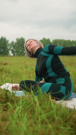 middle-aged woman with grey hair seated on yoga mat practicing side bend pose, hands stretched over her head, in a vast grassy field under cloudy skies, with trees lining the distance