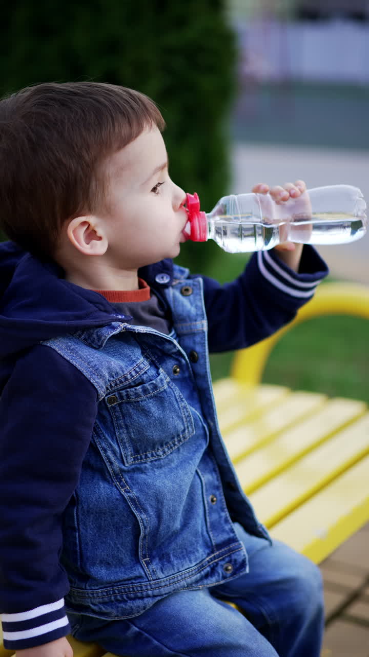 Pensive Caucasian toddler in jeans suit sits outdoors. Child drinks water from bottle. Vertical video.