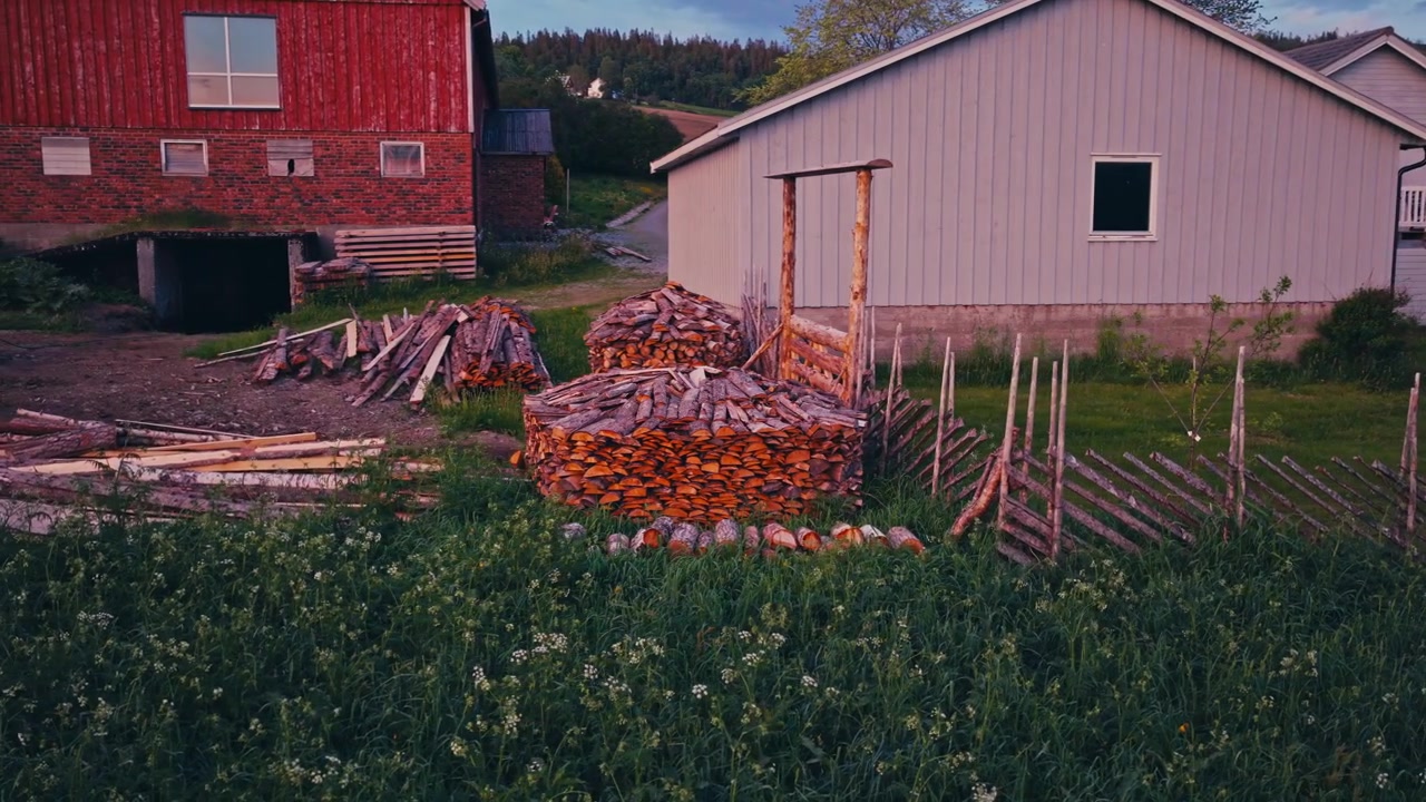 En haug med tømmerstokker står ved siden av en trelåve og et hus i et landlig landskap. Roterende droneopptak