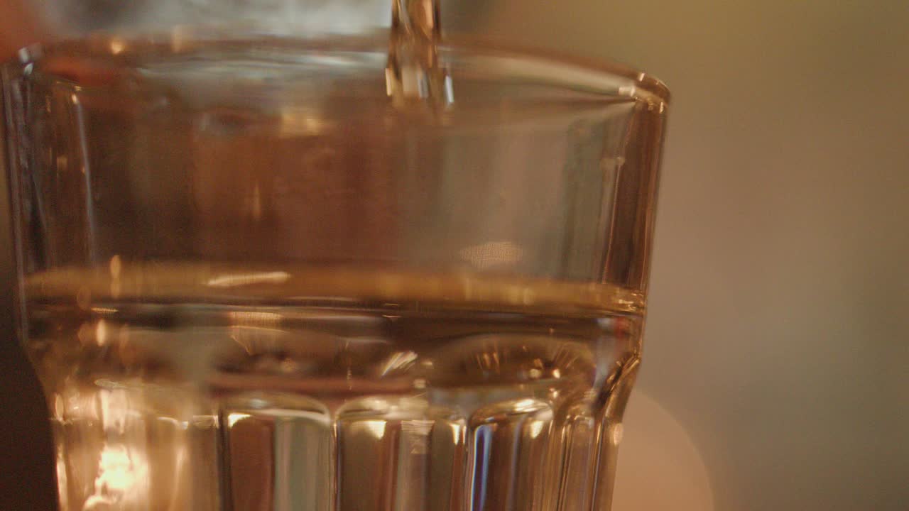 A close-up view of water being poured into a clear glass, with warm ambient lighting and a softly blurred restaurant background, captured in slow motion