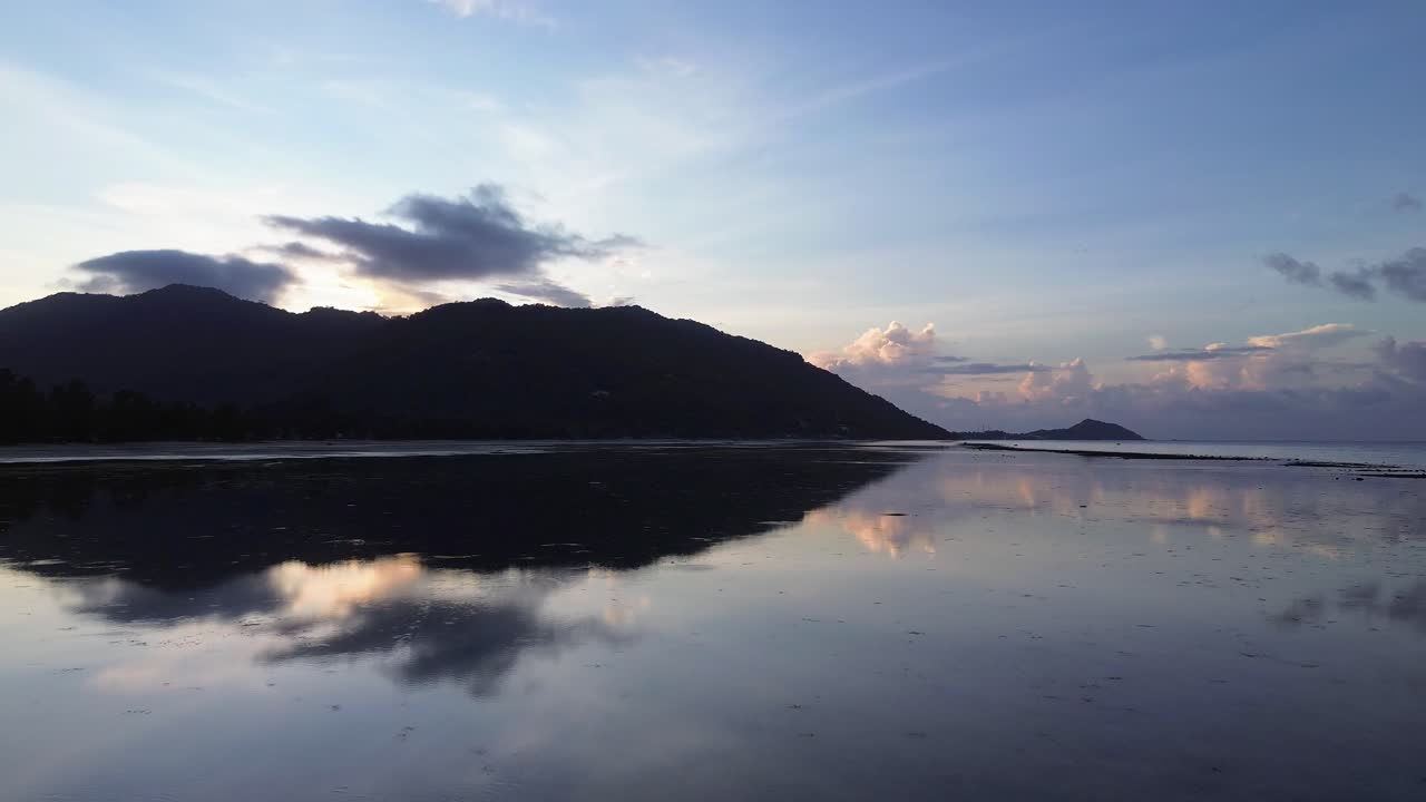 Drone footage capturing sunrise over Koh Phangan Thailand with the dark silhouette of a mountain and the reflection of it shimmering on the calm tropical sea