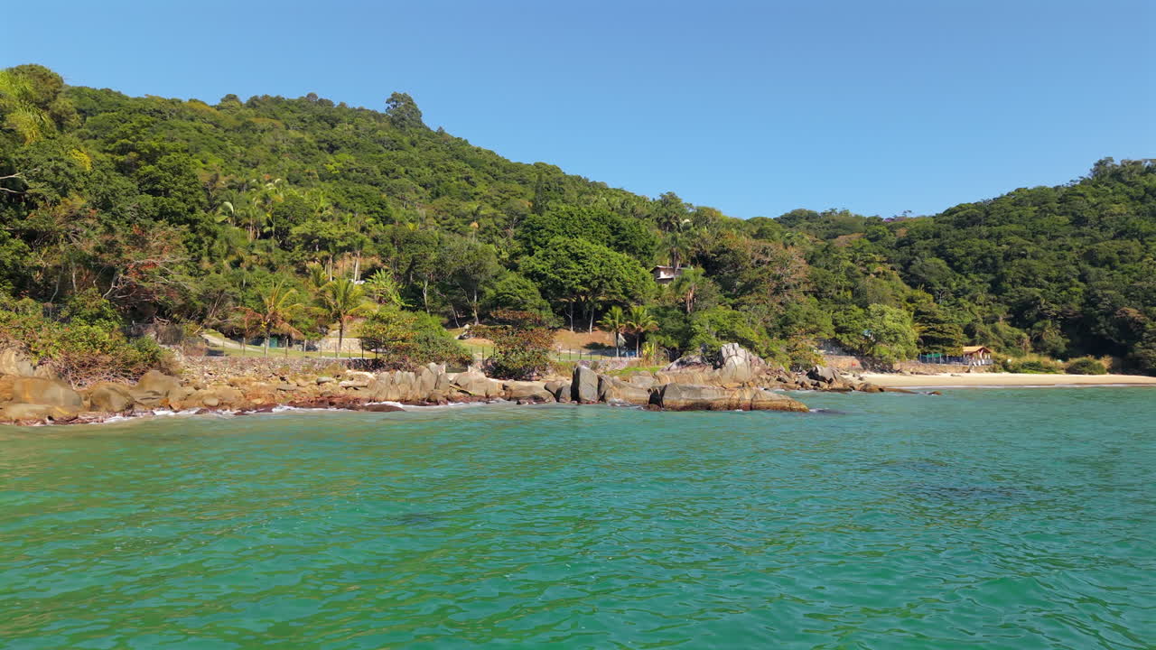 Drone shot at water level moving past a pier with birds, showing waves hitting rocks, vegetation, distant beach with houses and clear blue sky
