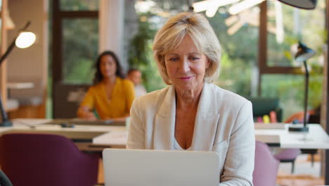 mature businesswoman working on laptop at desk in office pausing to look out of window