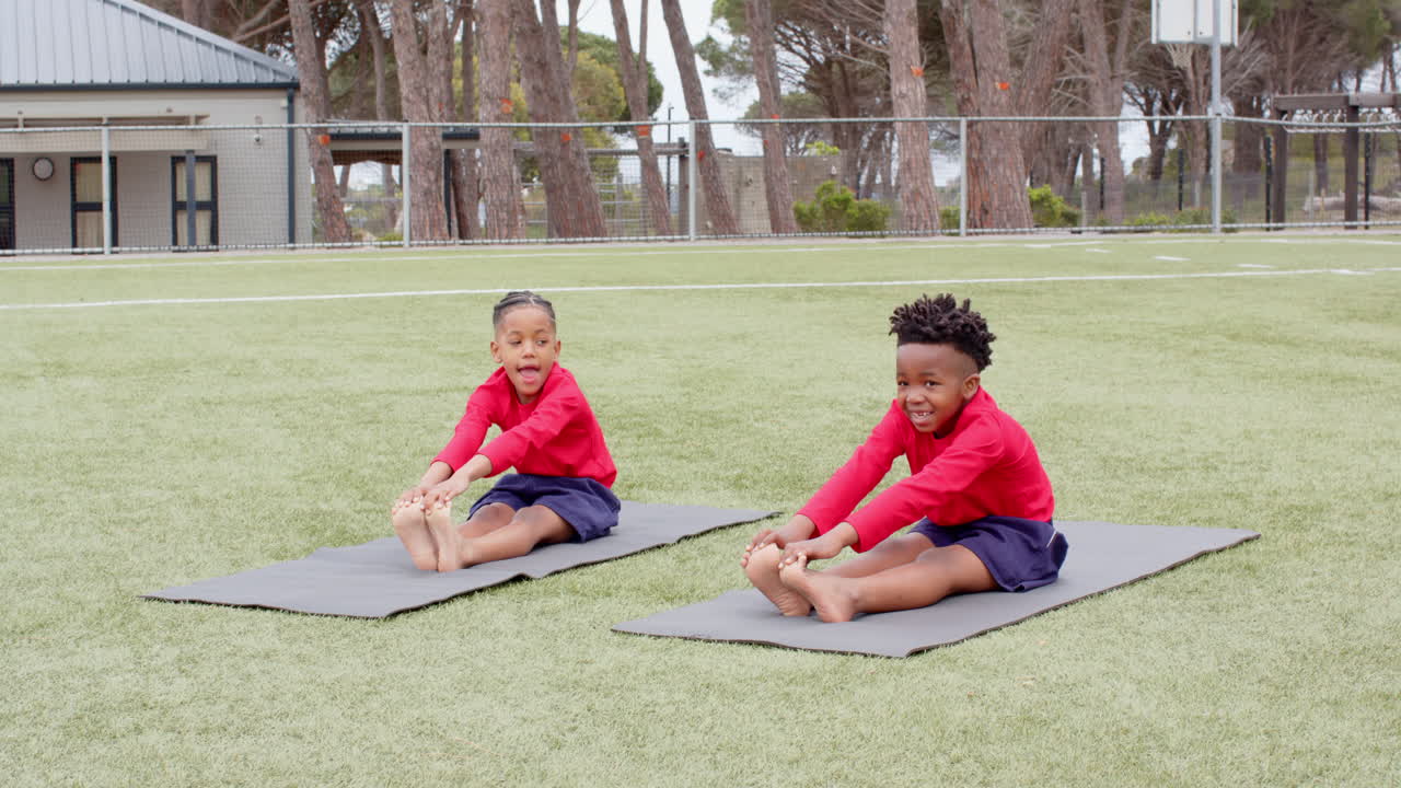 Multiracial boys stretching on yoga mats on school field, smiling and enjoying exercise at school