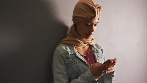 Asian-female-student-wearing-a-beige-hijab-leaning-against-a-wall-and-using-a-smartphone