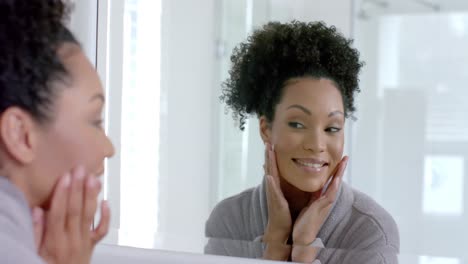 Happy-biracial-woman-in-bathrobe-looking-in-mirror-in-bathroom,-slow-motion