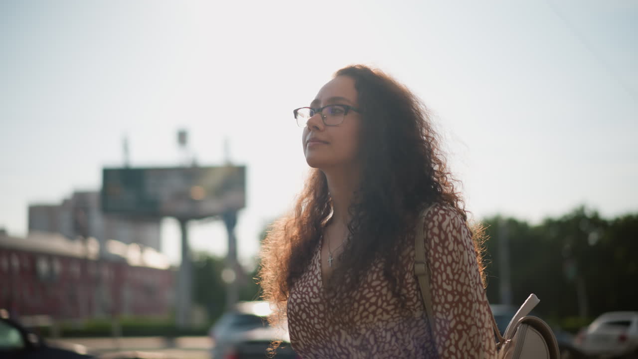 joven con gafas caminando con confianza en una ciudad concurrida, ajustando sus gafas mientras los coches pasan en el fondo, con edificios y la suave luz del sol iluminando su cara