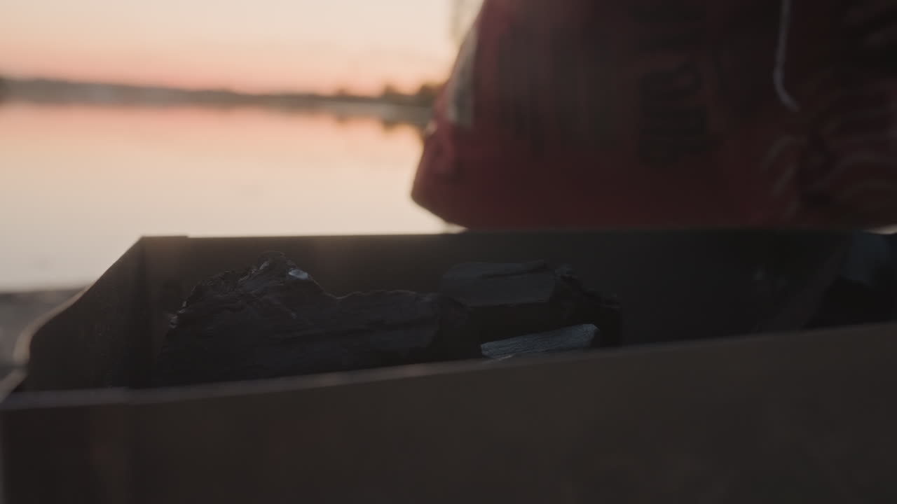 Mani che mettono il carbone nella griglia al tramonto in riva al lago, primo piano di scatola di cartone e bricchetti, dolci riflessi sull'acqua e crepuscolo caldo, dita che sistemano il combustibile per cucinare sul fuoco da campo, riva tranquilla