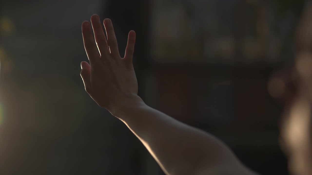 Una mano e un avambraccio umani sono sollevati contro uno sfondo interno fioco, con una morbida retroilluminazione che crea una silhouette netta, un ambiente circostante sfocato e un'atmosfera suggestiva.