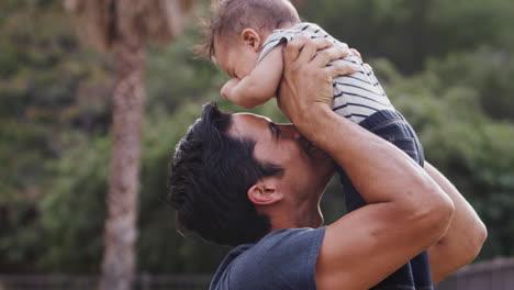 Millennial-Hispanic-man-lifting-his-baby-son-in-the-air-in-the-park,-close-up