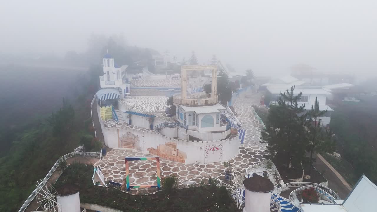 Drone view of a fog-covered Greek-style resort on a mountain ridge. Misty atmosphere and white-blue design create a cinematic, serene, and mysterious aesthetic