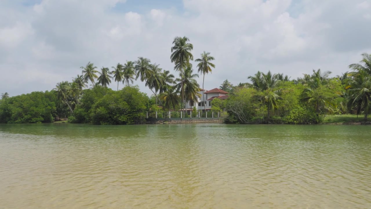 Ampia vista diurna del fiume Hikkaduwa in Sri Lanka con acqua increspata, palme da cocco e una villa sul fiume incorniciata da una lussureggiante vegetazione tropicale sotto un cielo nuvoloso.